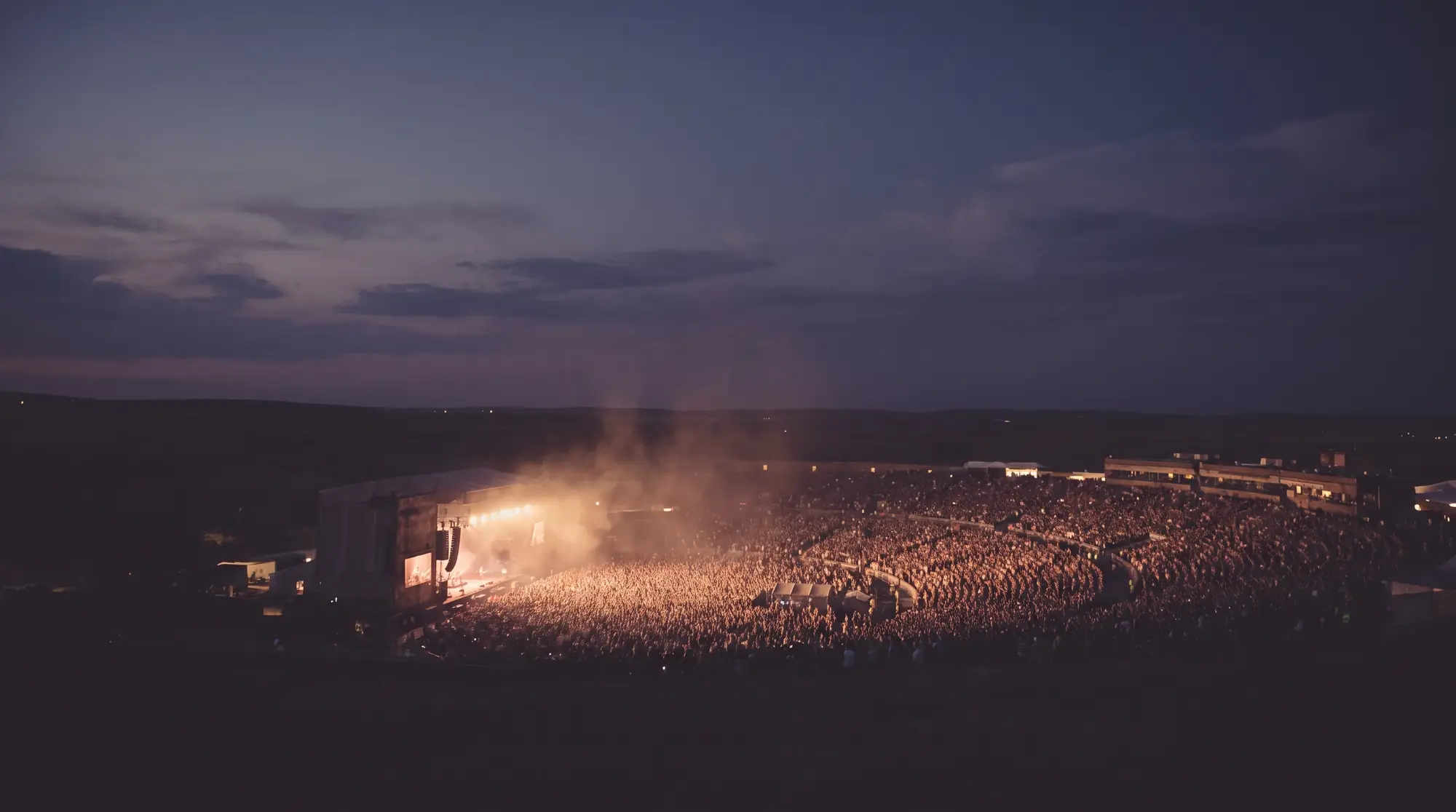 A large open-air concert at dusk, warm stage lights over a vast crowd